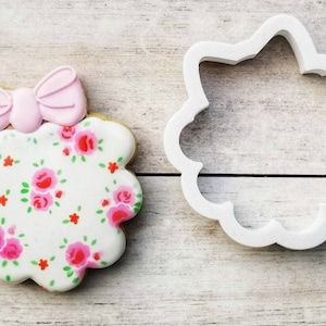 May include: A white cookie with a pink bow on top and a floral pattern. The cookie is next to a white plastic cookie cutter in the shape of a flower.