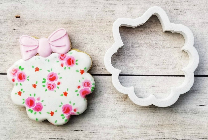 May include: A white cookie with a pink bow on top and a floral pattern. The cookie is next to a white plastic cookie cutter in the shape of a flower.