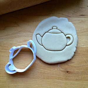 May include: A white plastic cookie cutter in the shape of a teapot. The cutter is on a wooden surface next to a cookie that has been cut out with the cutter.