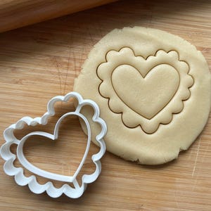 May include: A white plastic heart-shaped cookie cutter with a scalloped edge. The cutter is placed on top of a plain heart-shaped cookie dough.