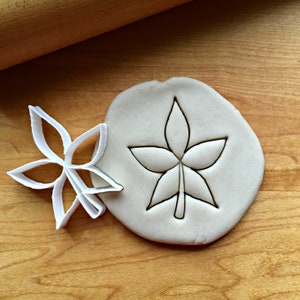 May include: A white plastic flower-shaped cookie cutter next to a piece of dough with a flower imprint. The cutter is on a wooden surface with a rolling pin in the background. The dough has a flower shape pressed into it.