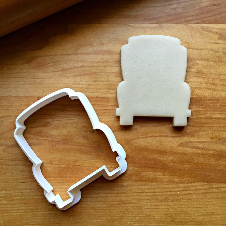 May include: A white plastic cookie cutter in the shape of a truck is next to a truck-shaped cookie on a wooden surface. The cookie cutter is empty, and the cookie is beige. A rolling pin is in the background.