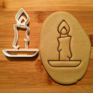 May include: A white plastic cookie cutter shaped like a candle with a flame on top. The cutter is on a wooden surface next to a brown cookie dough cutout of the same candle shape.
