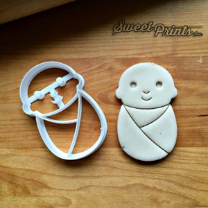 May include: A white plastic cookie cutter in the shape of a baby swaddled in a blanket. Next to it is a cookie made from dough, also in the shape of a swaddled baby.