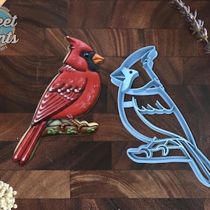 May include: A red cardinal bird cookie decorated with icing next to a light blue plastic cardinal bird cookie cutter. The cookie features a black face mask, orange beak, and brown branch detail. The text Sweet Prints Established 2015 is visible.