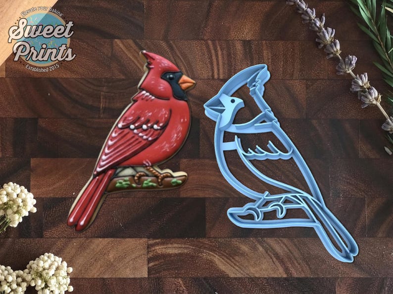 May include: A red cardinal bird cookie decorated with icing next to a light blue plastic cardinal bird cookie cutter. The cookie features a black face mask, orange beak, and brown branch detail. The text Sweet Prints Established 2015 is visible.