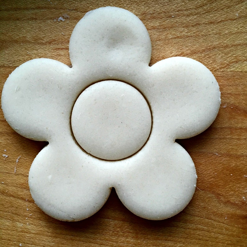 Puede incluir: Un cortador de galletas blanco en forma de flor con un corte redondo en el centro. El cortador est&aacute; sobre una superficie de madera.