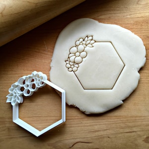 May include: White plastic cookie cutter in the shape of a hexagon with a floral design on one side. The cutter is on a wooden surface with a piece of dough that has been cut out with the cutter.
