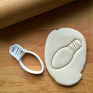 May include: White plastic cookie cutter shaped like a light bulb. The cutter is next to a piece of cookie dough with the light bulb shape cut out.