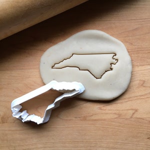 May include: A white plastic cookie cutter in the shape of the state of North Carolina. The cutter is on a wooden surface next to a piece of cookie dough.
