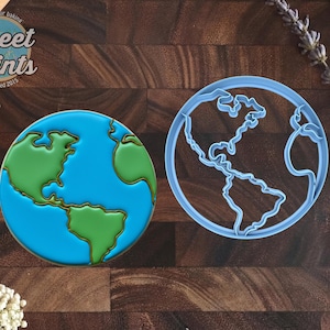 May include: A round cookie decorated with green continents and blue oceans alongside a light blue plastic globe-shaped cookie cutter. The cookie features detailed outlines of North and South America, matching the shape of the cutter.
