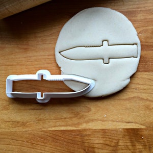 May include: A white plastic knife-shaped cookie cutter next to a circle of unbaked dough with a knife imprint. The cutter has a serrated edge and a handle. The dough is on a wooden surface.