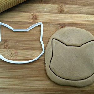 May include: A white plastic cat-shaped cookie cutter on a wooden surface next to a brown cookie dough cut out in the shape of a cat.