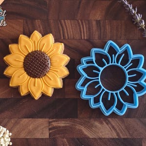 May include: A sunflower-shaped cookie decorated with yellow icing and a brown center, positioned next to a matching blue plastic sunflower cookie cutter. The cookie features detailed petal lines and a cross-hatched center design.