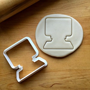 May include: White plastic cookie cutter shaped like a computer monitor. The cutter is on a wooden surface with a piece of dough that has been cut out with the cutter.