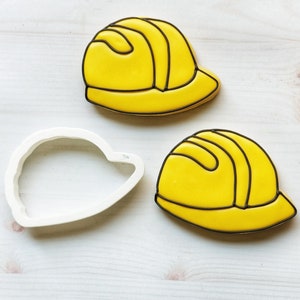 May include: Two yellow construction hard hat cookies with black outlines and a white cookie cutter. The cookies are decorated with yellow icing and black details. The cookie cutter is white and shaped like a hard hat.