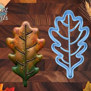 May include: A light blue plastic oak leaf cookie cutter next to a decorated cookie shaped like an oak leaf with a gradient of green, orange, and brown icing, featuring a dark brown piped vein and small gold sprinkles along the center stem.