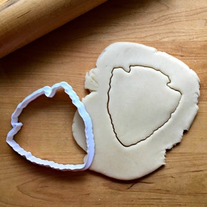 May include: A white plastic cookie cutter in the shape of a pointed arrow, used to cut out a cookie from a sheet of unbaked dough.