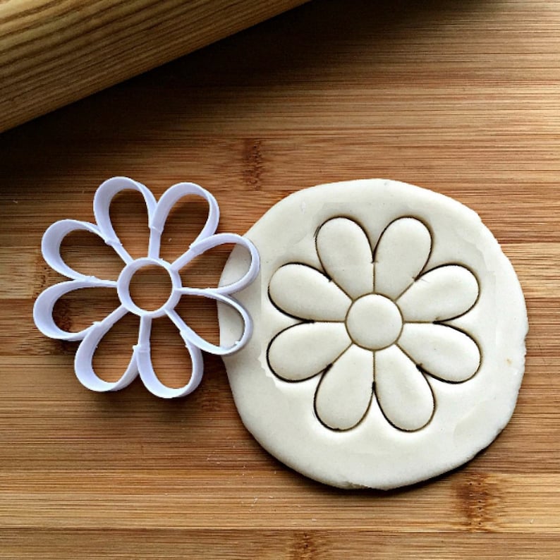 May include: A white daisy-shaped cookie cutter next to a round of dough with a daisy imprint. The cutter is made of plastic and has a simple, clean design. The dough is on a wooden surface.