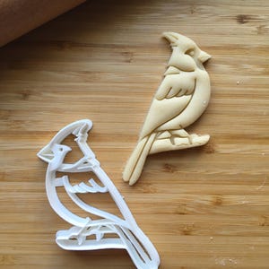 May include: A white plastic cardinal bird cookie cutter next to a matching raw dough cutout of a cardinal bird perched on a branch. The cookie cutter features a detailed outline of the bird with a crest, wing, and tail feathers clearly defined.