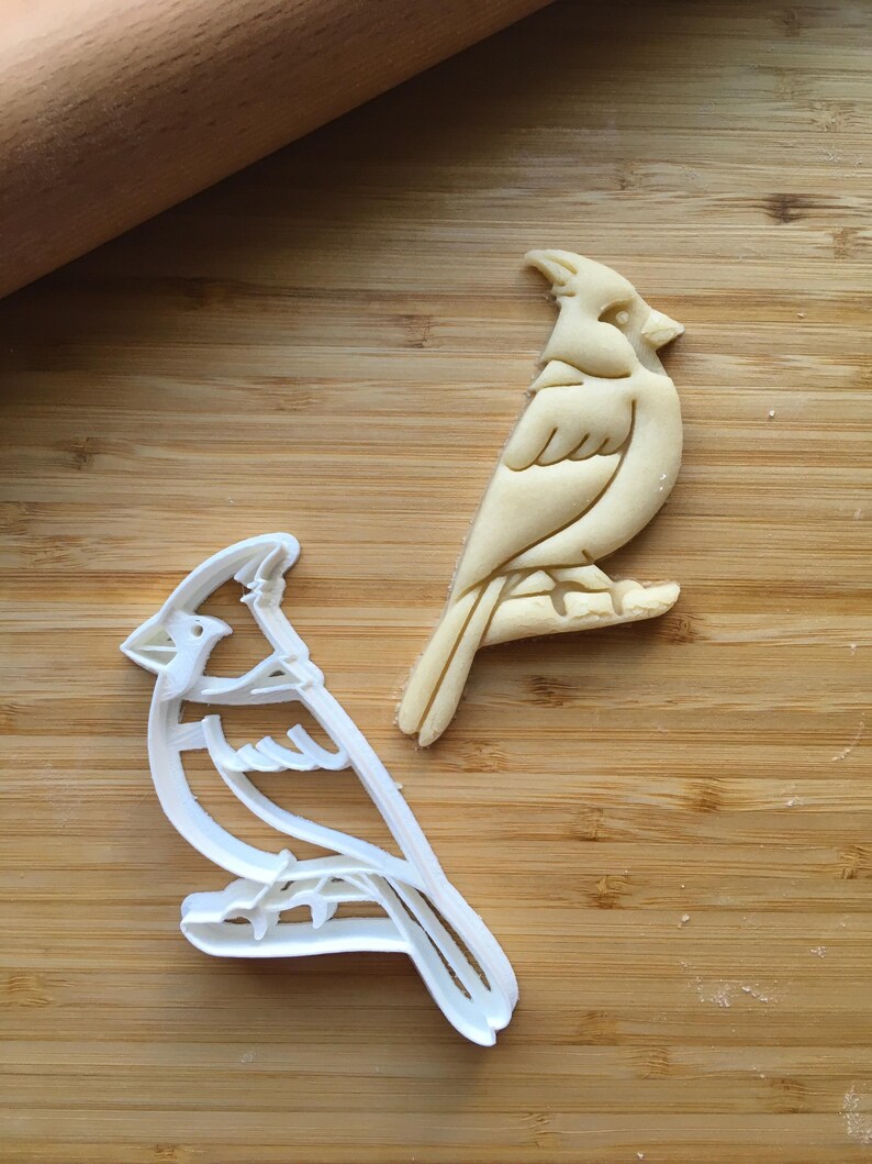 May include: A white plastic cardinal bird cookie cutter next to a matching raw dough cutout of a cardinal bird perched on a branch. The cookie cutter features a detailed outline of the bird with a crest, wing, and tail feathers clearly defined.