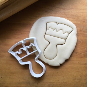 May include: A white plastic cookie cutter in the shape of a paintbrush next to a piece of dough with a paintbrush imprint. The dough is on a wooden surface, with a rolling pin visible in the background. The cookie cutter is a craft supply.