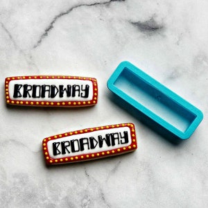 May include: Two red and white cookies decorated with black icing that reads "BROADWAY" surrounded by yellow dots. A blue plastic cookie cutter in the shape of a rectangle is also shown.