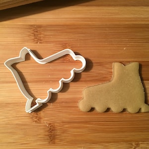 May include: White plastic roller skate cookie cutter on a wooden surface next to a brown sugar cookie in the shape of a roller skate.
