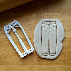 May include: A white plastic cookie cutter shaped like a pair of jeans. The cutter is shown next to a cookie that has been cut out using the cutter.