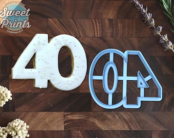 May include: A number 40 cookie decorated with light blue icing and small white sprinkles, positioned next to a light blue plastic number 40 cookie cutter. The cookie and cutter are displayed on a dark brown wooden surface with floral accents.
