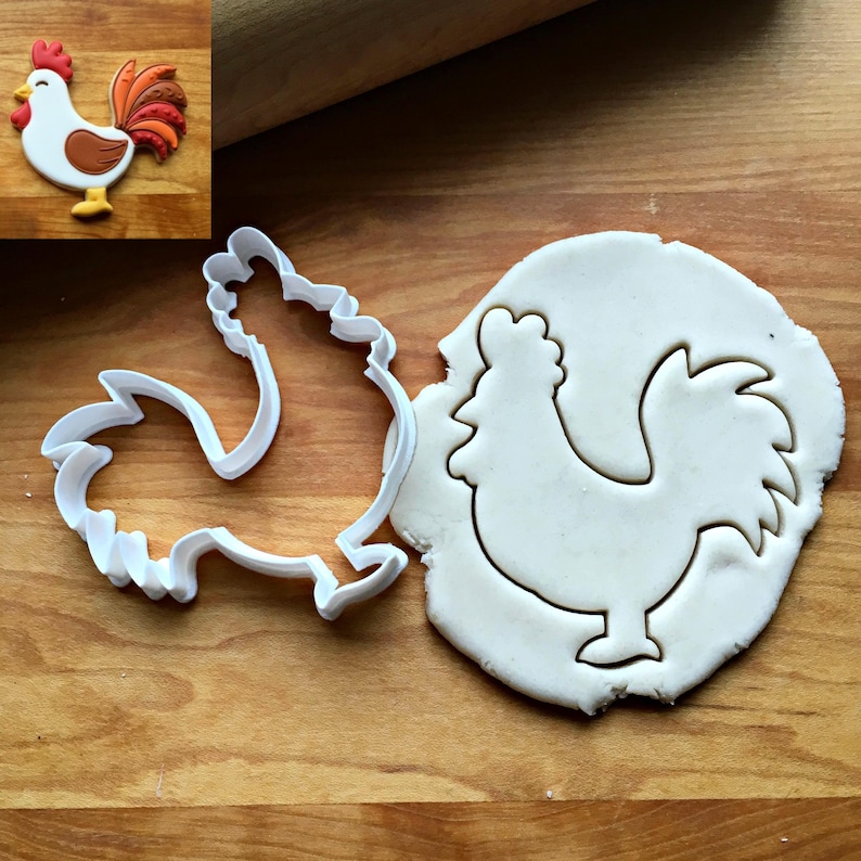 May include: A white plastic rooster-shaped cookie cutter next to a piece of rolled-out white dough with a rooster-shaped cutout. An inset image shows a finished cookie decorated with white, brown, orange, and red icing in the shape of a rooster.