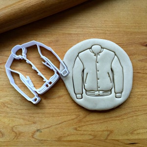 May include: White plastic cookie cutter in the shape of a bomber jacket. The cutter is next to a piece of unbaked cookie dough that has been pressed with the cutter.