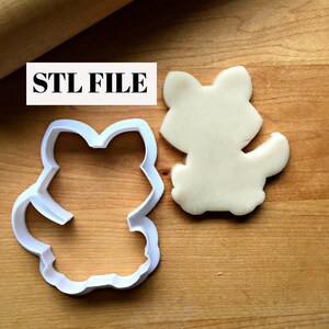 May include: A white plastic cookie cutter in the shape of a fox, next to a fox-shaped cookie. The text "STL FILE" is in a white rectangle. The objects are on a wooden surface.