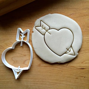 May include: White plastic cookie cutter shaped like a heart with an arrow through it. The cutter is on a wooden surface next to a cookie that has been cut with the cutter.