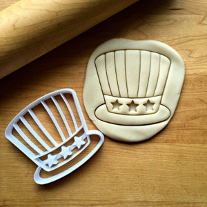 May include: A white plastic cookie cutter in the shape of a red, white, and blue Uncle Sam hat. The cookie cutter is on a wooden surface next to a cookie dough cutout of the same shape.