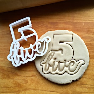 May include: White plastic cookie cutter in the shape of the number 5 with the word "five" written in a cursive font. The cookie cutter is next to a cookie dough circle with the number 5 and the word "five" cut out.