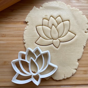 May include: A white plastic lotus flower cookie cutter next to a lotus flower cut-out in dough. The dough is on a wooden cutting board with a rolling pin in the background. The cookie cutter is a detailed outline of the flower.