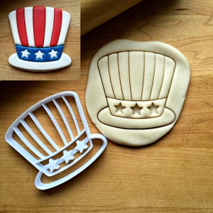 May include: A white plastic cookie cutter shaped like a top hat with vertical stripes and three stars on the brim. Beside it, a piece of raw cookie dough is pressed with the same hat design, and a finished cookie shows red, white, and blue icing.