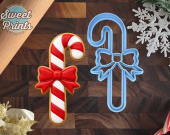 May include: A candy cane cookie with red and white stripes and a red bow, alongside a light blue plastic candy cane cookie cutter with a bow shape. The cookie and cutter are displayed on a dark wood surface with a snowflake and text.
