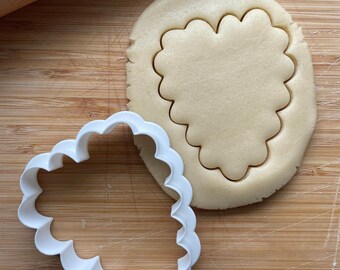 May include: A white plastic cookie cutter shaped like a heart with scalloped edges, on top of a round piece of cookie dough with a heart-shaped cutout.