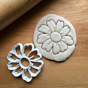 May include: A white plastic cookie cutter in the shape of a flower with eight petals. The cookie cutter is on a wooden surface next to a cookie dough cutout of the same flower shape.