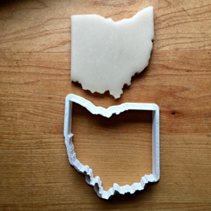 May include: White plastic cookie cutter in the shape of the state of Ohio. The cutter is on a wooden surface.