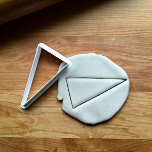 May include: A white plastic triangle cookie cutter on a wooden surface with a piece of dough that has been cut out with the cutter.