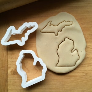May include: Two white plastic cookie cutters shaped like the state of Michigan. The larger cutter is of the entire state, while the smaller cutter is of the lower peninsula. The cutters are on a wooden surface with a piece of dough that has been cut out with the larger cutter.