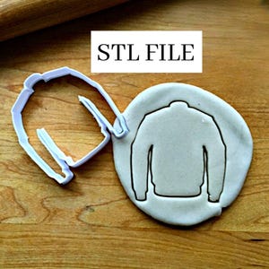 Może przedstawiać: Biała plastikowa wykrawaczka do ciastek w kształcie swetra, obok ciastka z odciśniętym konturem swetra. Tekst "STL FILE" znajduje się w białym prostokącie powyżej.
