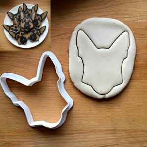 May include: A white plastic cookie cutter shaped like a German Shepherd dog head, shown next to a piece of rolled dough with the same shape cut out, and a small inset image of several finished cookies decorated to look like the dog breed.