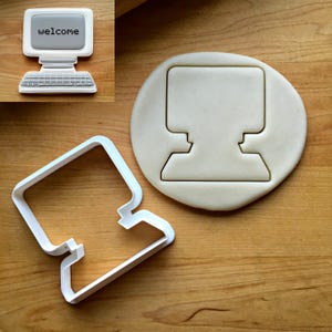 May include: A white plastic cookie cutter shaped like a vintage desktop computer, accompanied by a piece of dough cut into the same computer shape. The dough features a gray screen with the word welcome printed in black pixelated text.