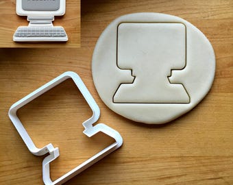 May include: A white plastic cookie cutter shaped like a vintage desktop computer, accompanied by a piece of dough cut into the same computer shape. The dough features a gray screen with the word welcome printed in black pixelated text.