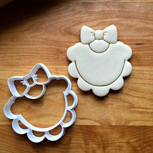 May include: A white plastic cookie cutter shaped like a baby bib with a bow on top. The cookie cutter is on a wooden surface.
