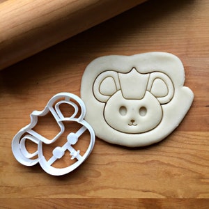 May include: A white plastic cookie cutter shaped like a cartoon mouse wearing a crown. The cutter is next to a cookie dough cutout of the same design.